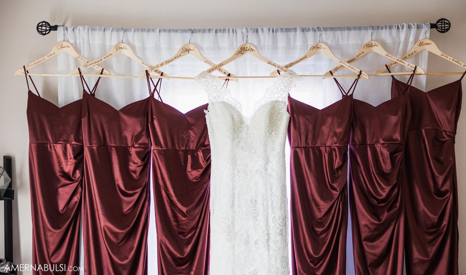Wedding dress and burgundy bridesmaid dresses hanging on display rod against white wall.