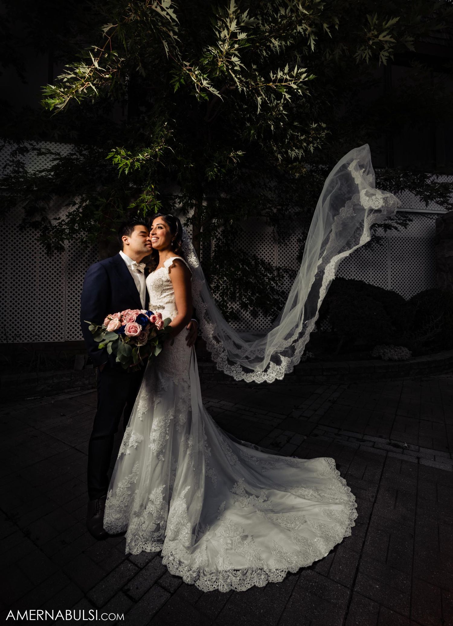 Dramatic evening wedding photo with bride's veil flowing in wind against dark background.