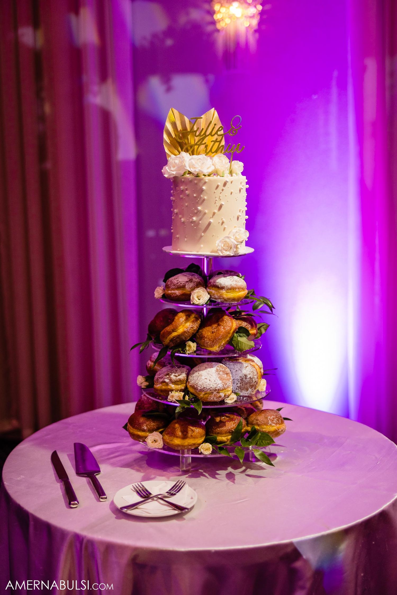 Elegant wedding dessert tower with white cake topped with gold crown and multiple tiers of treats in purple lighting.