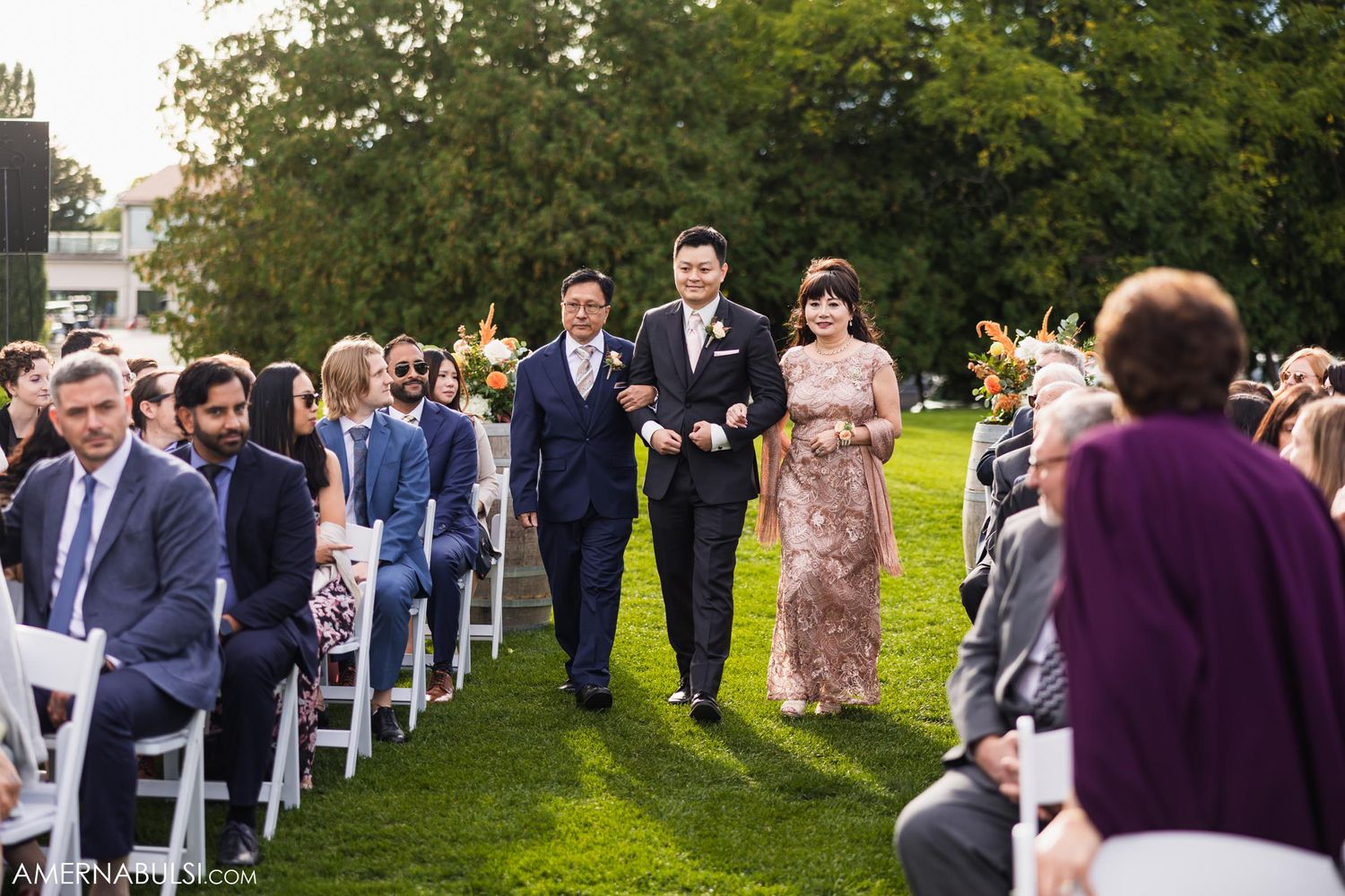Wedding party members walk down an outdoor aisle lined with white chairs and guests.
