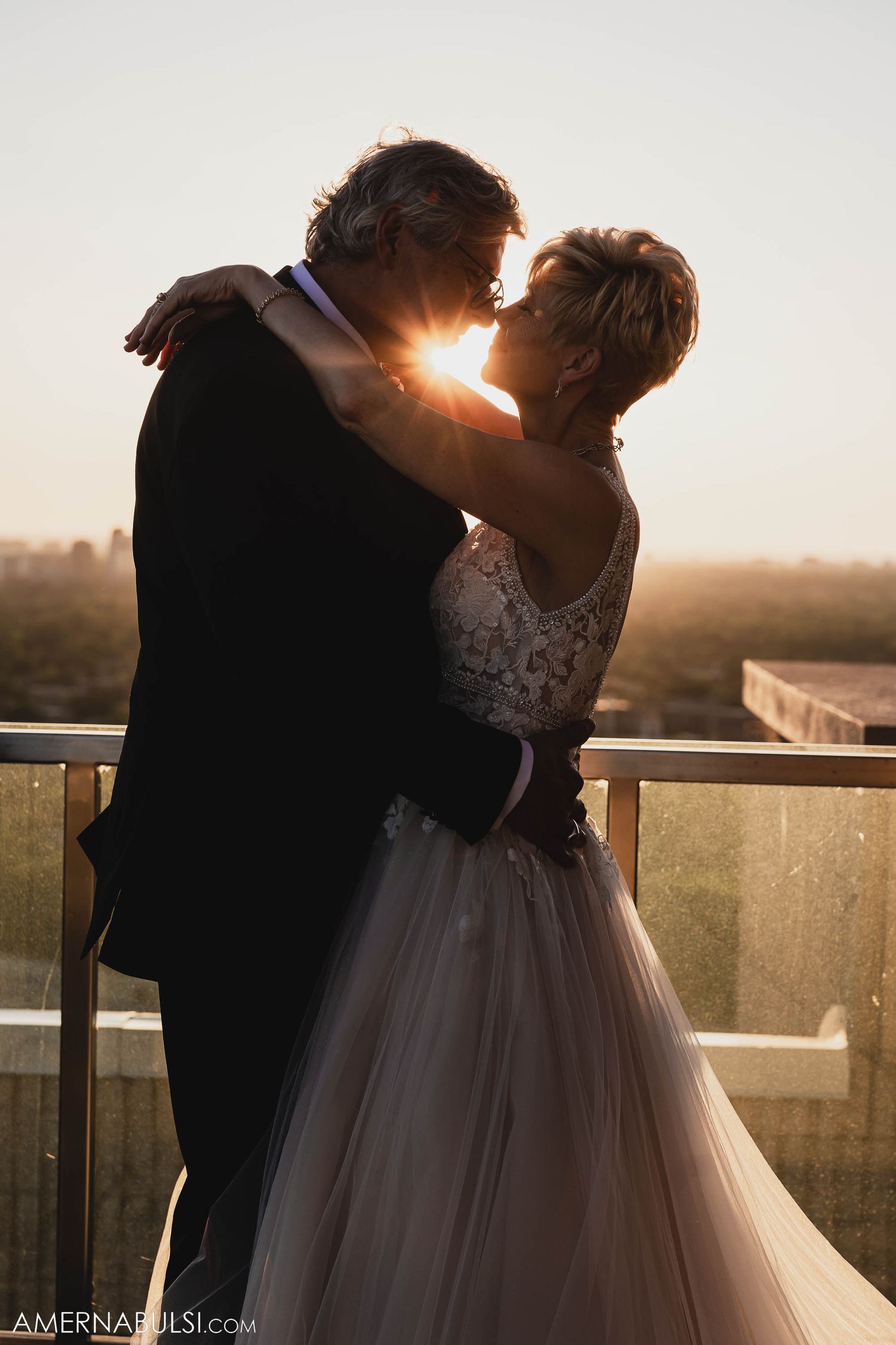 A couple embraces in formal wedding attire on a balcony during golden hour sunset.