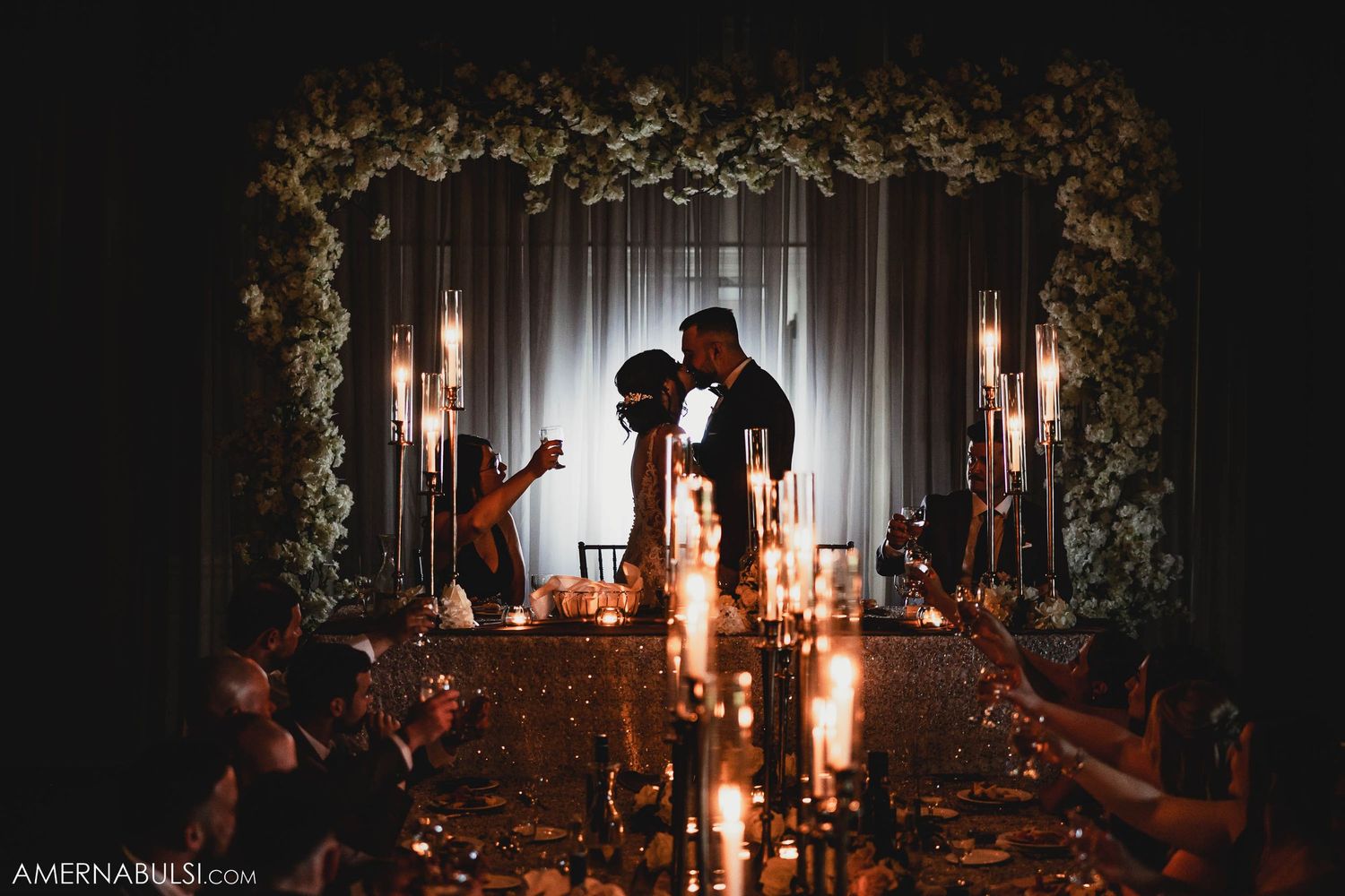 Silhouettes against a candlelit backdrop with floral arch during an elegant evening wedding ceremony.