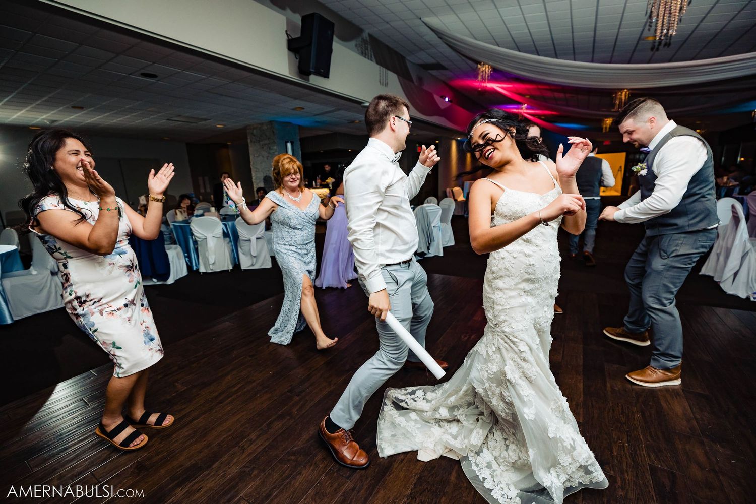 Wedding reception dance party with guests celebrating on wooden dance floor under blue lighting.