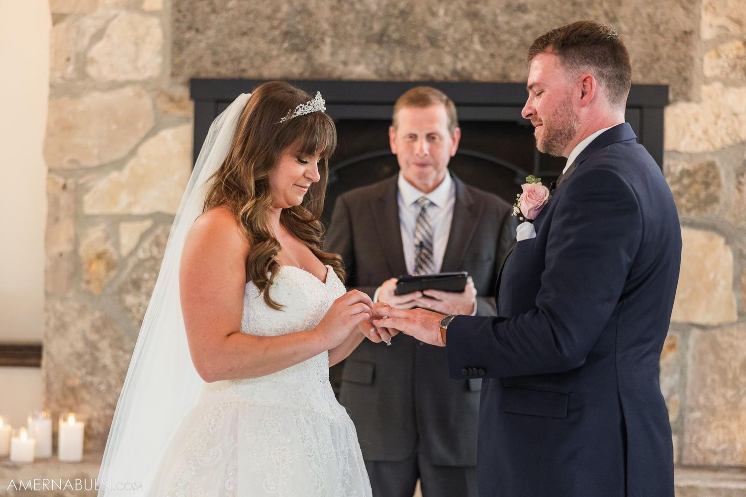 Intimate moment during ring exchange at wedding ceremony in front of stone fireplace.