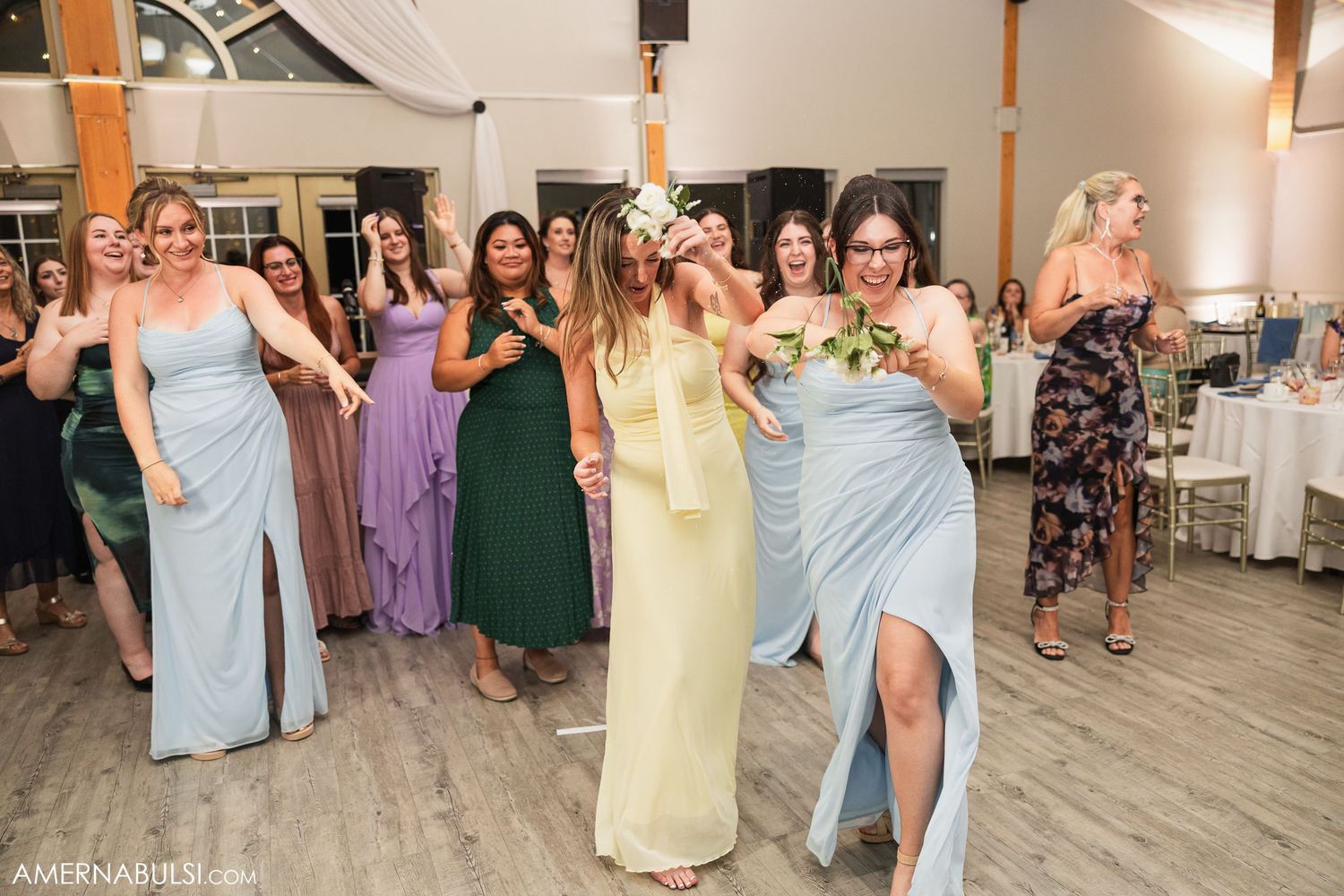 Wedding party and guests dancing together on a light wooden floor at an indoor reception venue.
