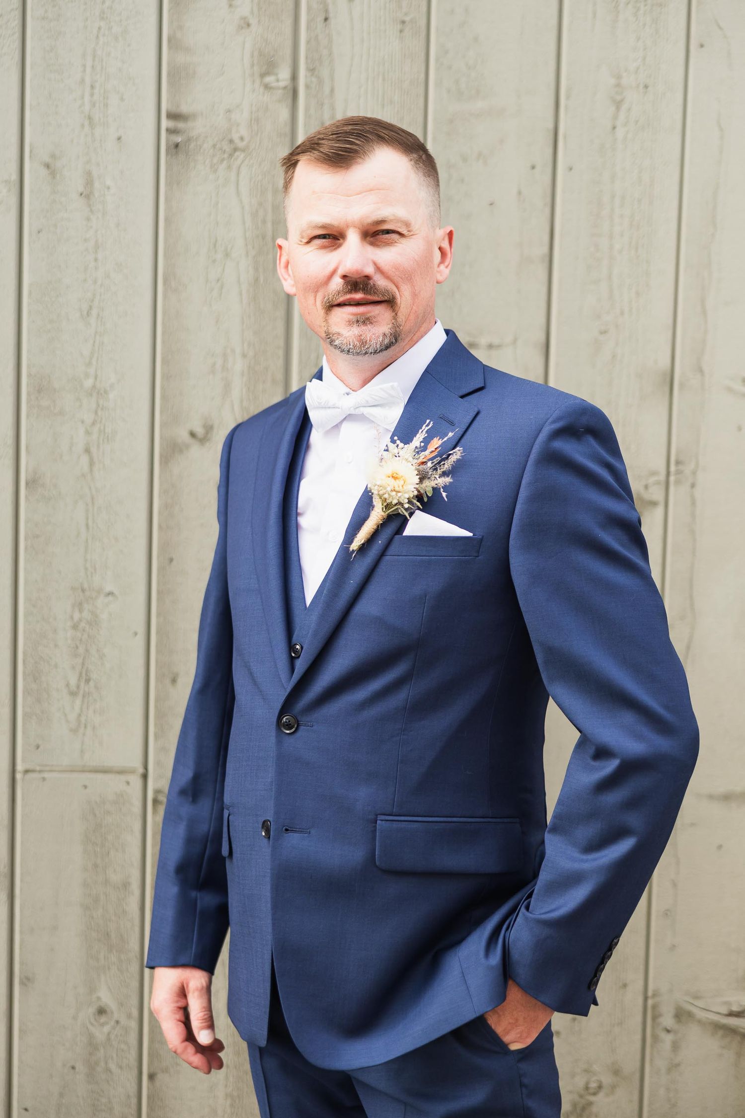 Groom poses against wooden wall wearing navy suit with boutonniere.