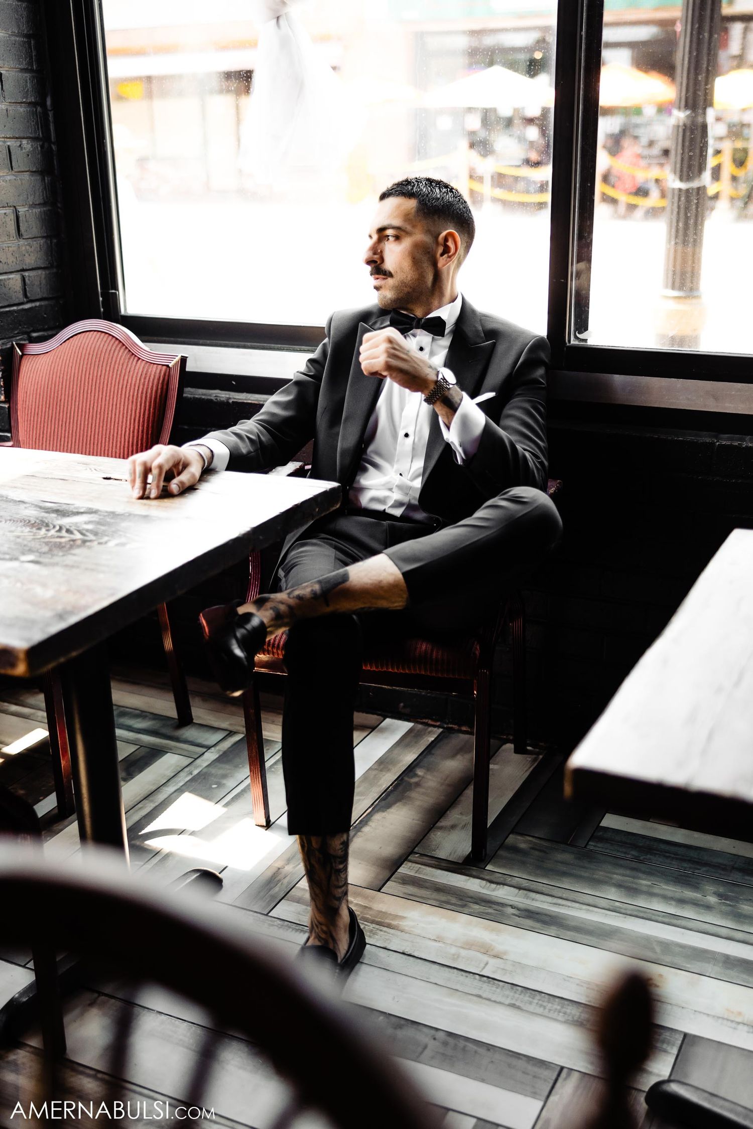 Person in formal attire seated at restaurant table near window with drink.
