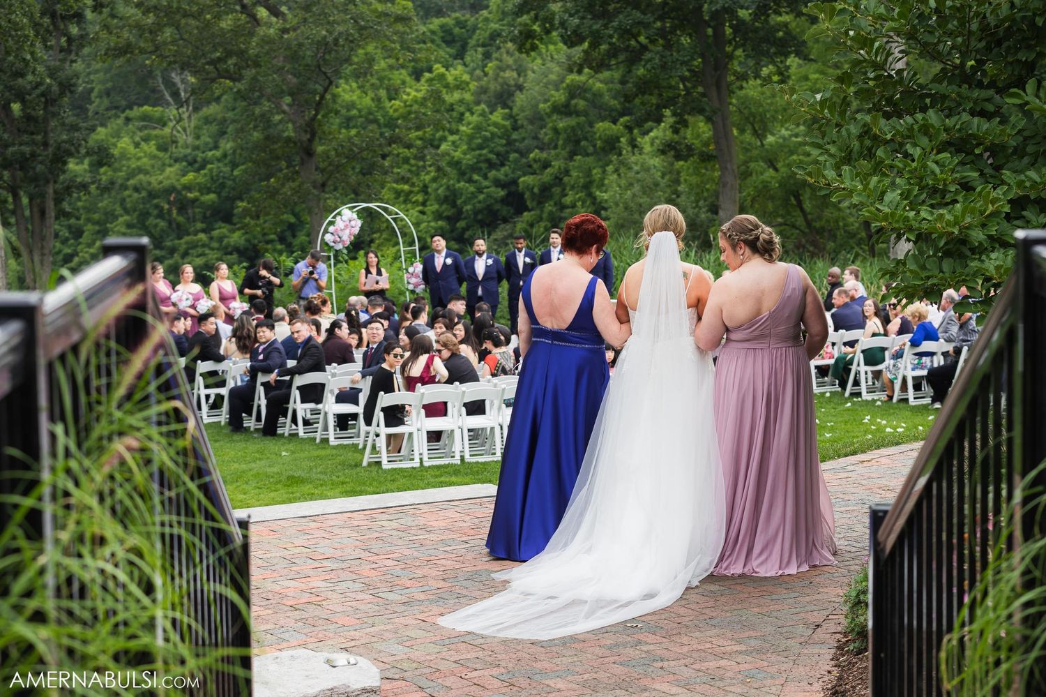 Wedding party members stand on brick pathway overlooking seated guests during outdoor ceremony.
