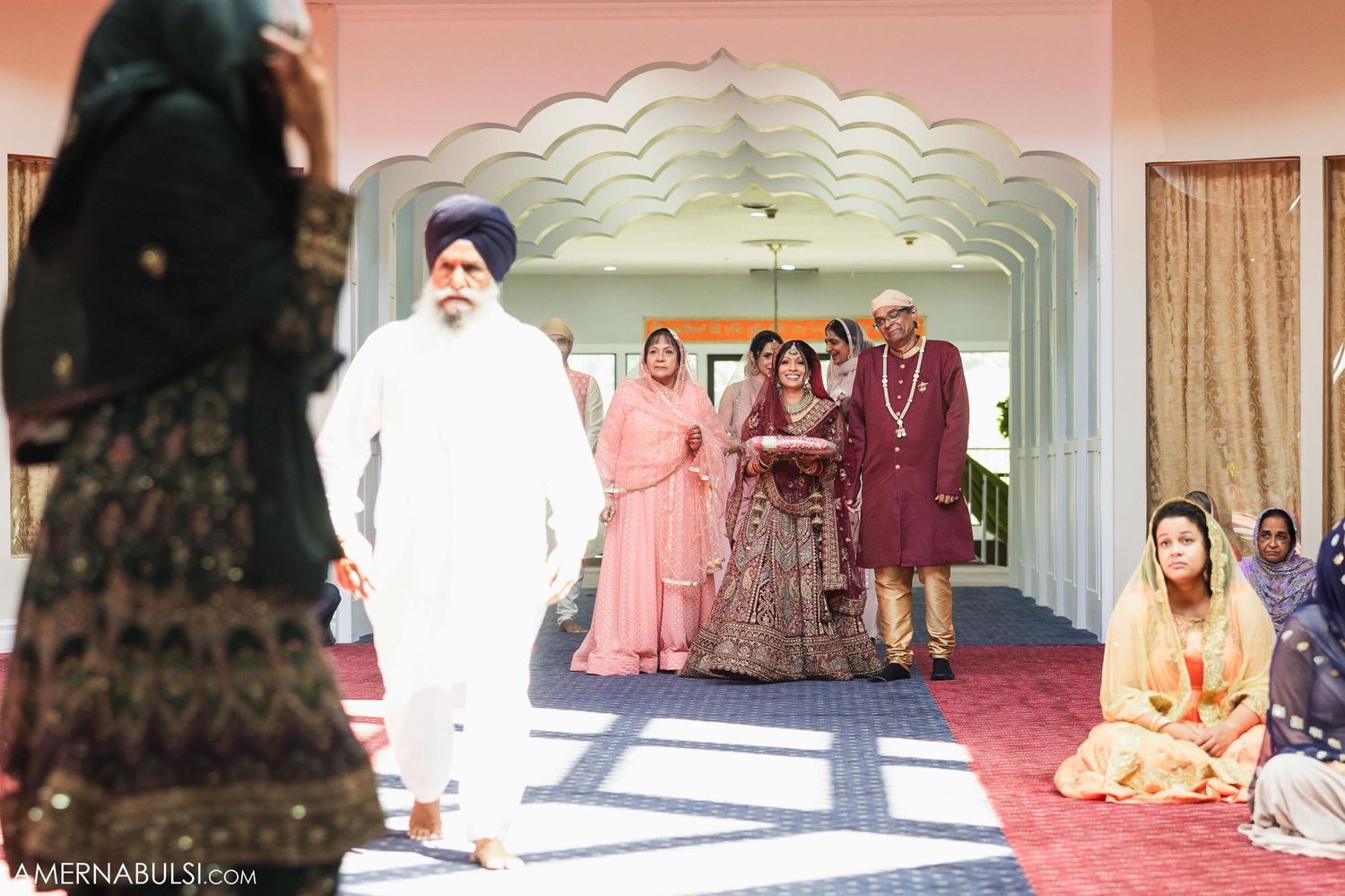 Traditional Indian wedding ceremony in a pink-walled room with decorative archway and gathered guests.