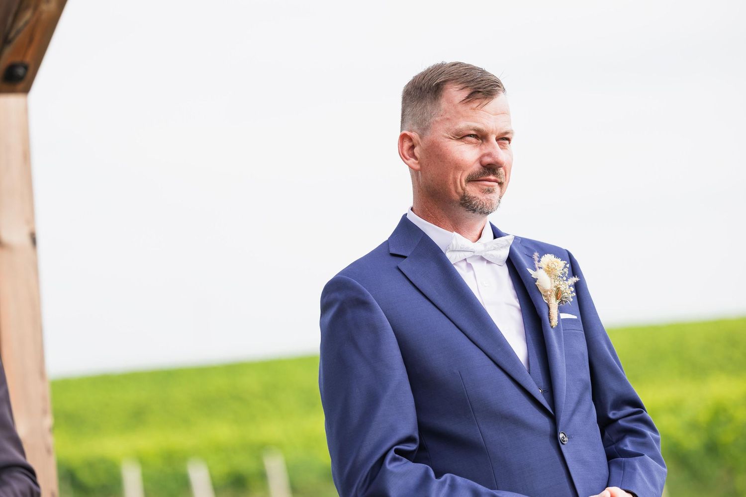 Groom in tailored navy suit waits at outdoor ceremony site.
