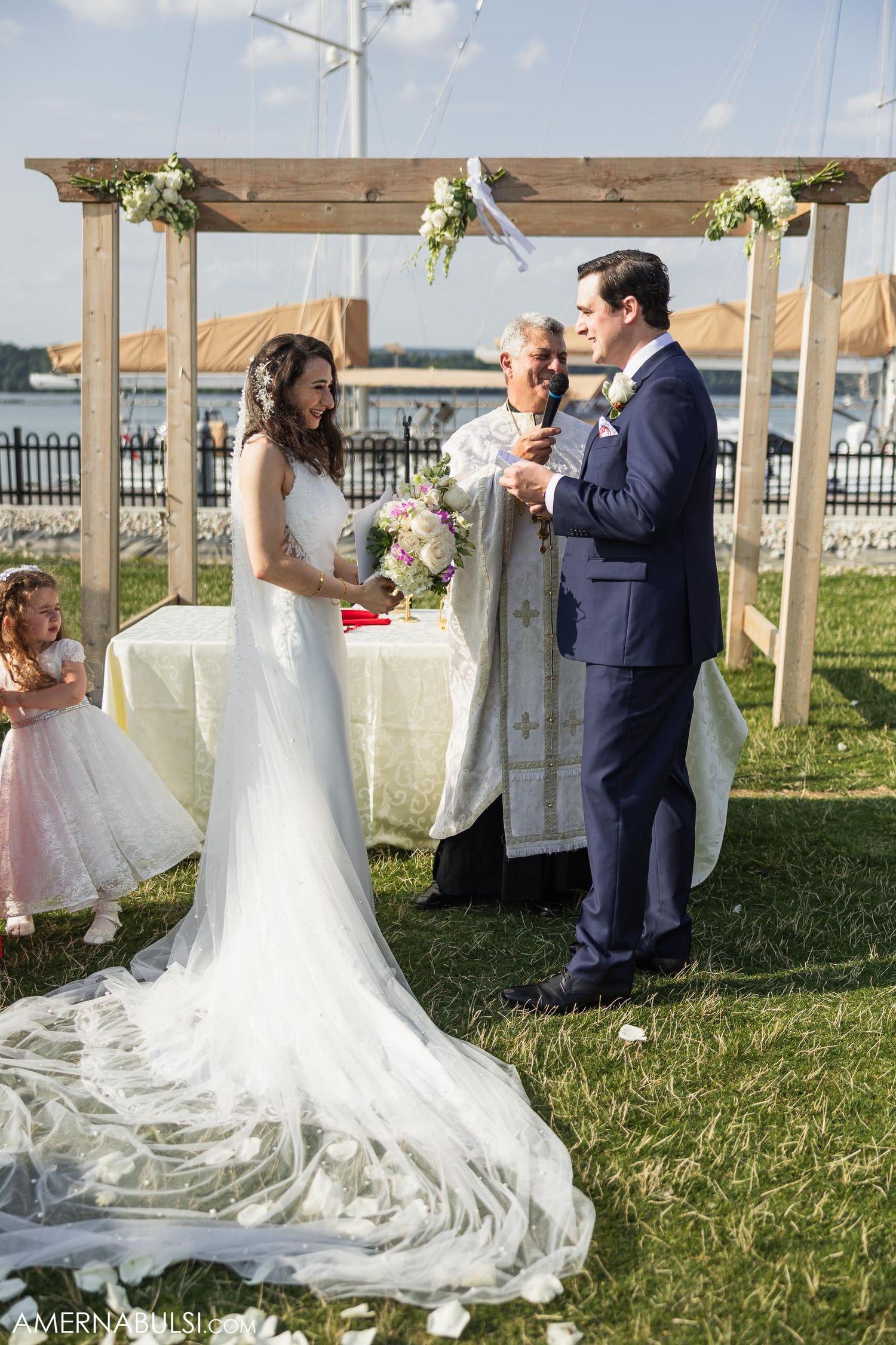 Wedding ceremony under wooden pergola decorated with white flowers at waterfront location.