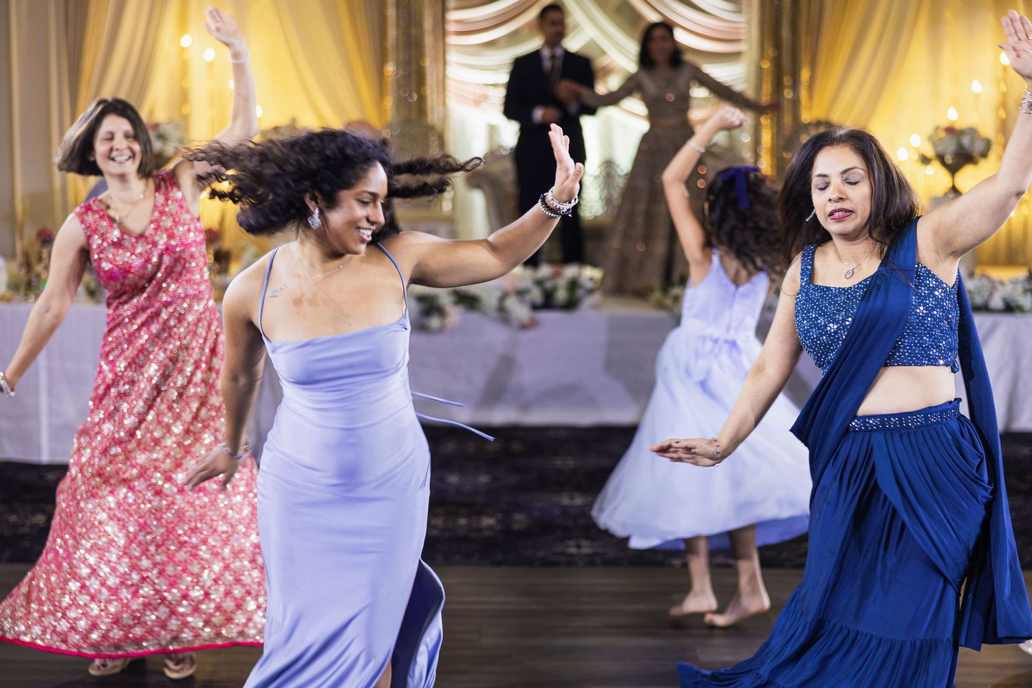 Wedding guests dancing joyfully in colorful dresses at an elegant reception with golden drapes.