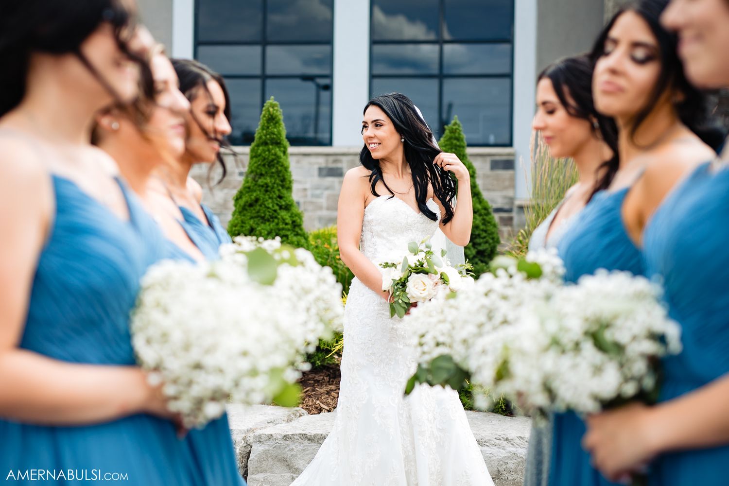Wedding party in blue dresses posing with white floral bouquets outside modern building.