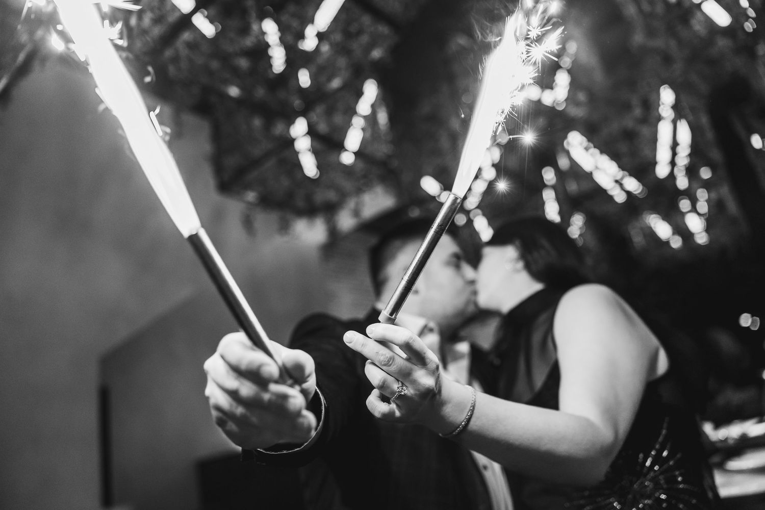 Black and white photo of couple kissing while holding sparklers at a celebration event.