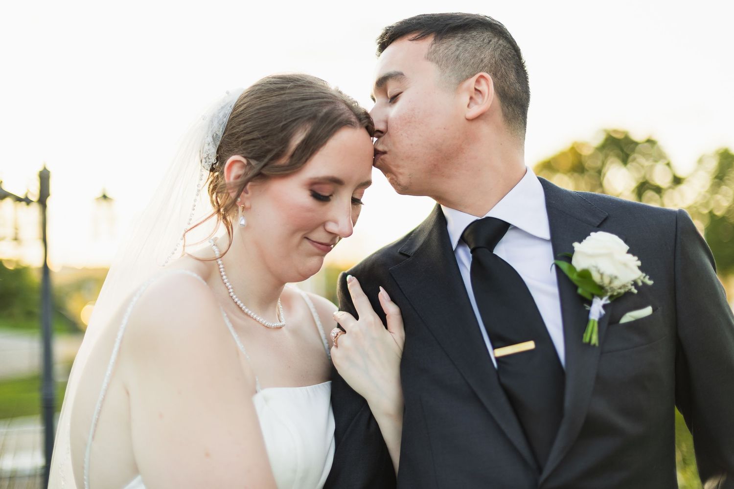 Groom kissing bride on forehead outdoors at golden hour during wedding portrait session.