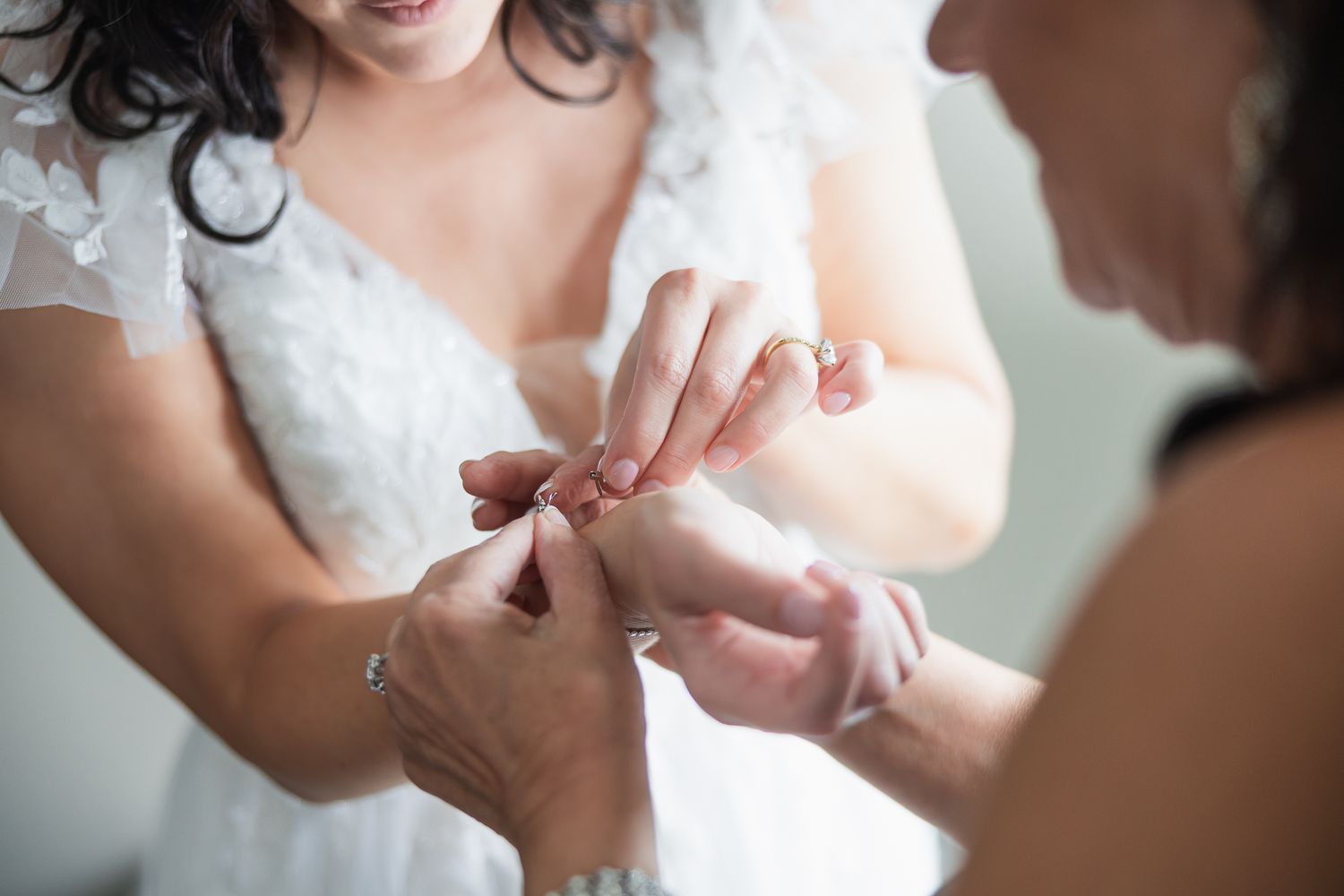 Mother helping bride put on bracelet while wearing lace wedding gown before ceremony.