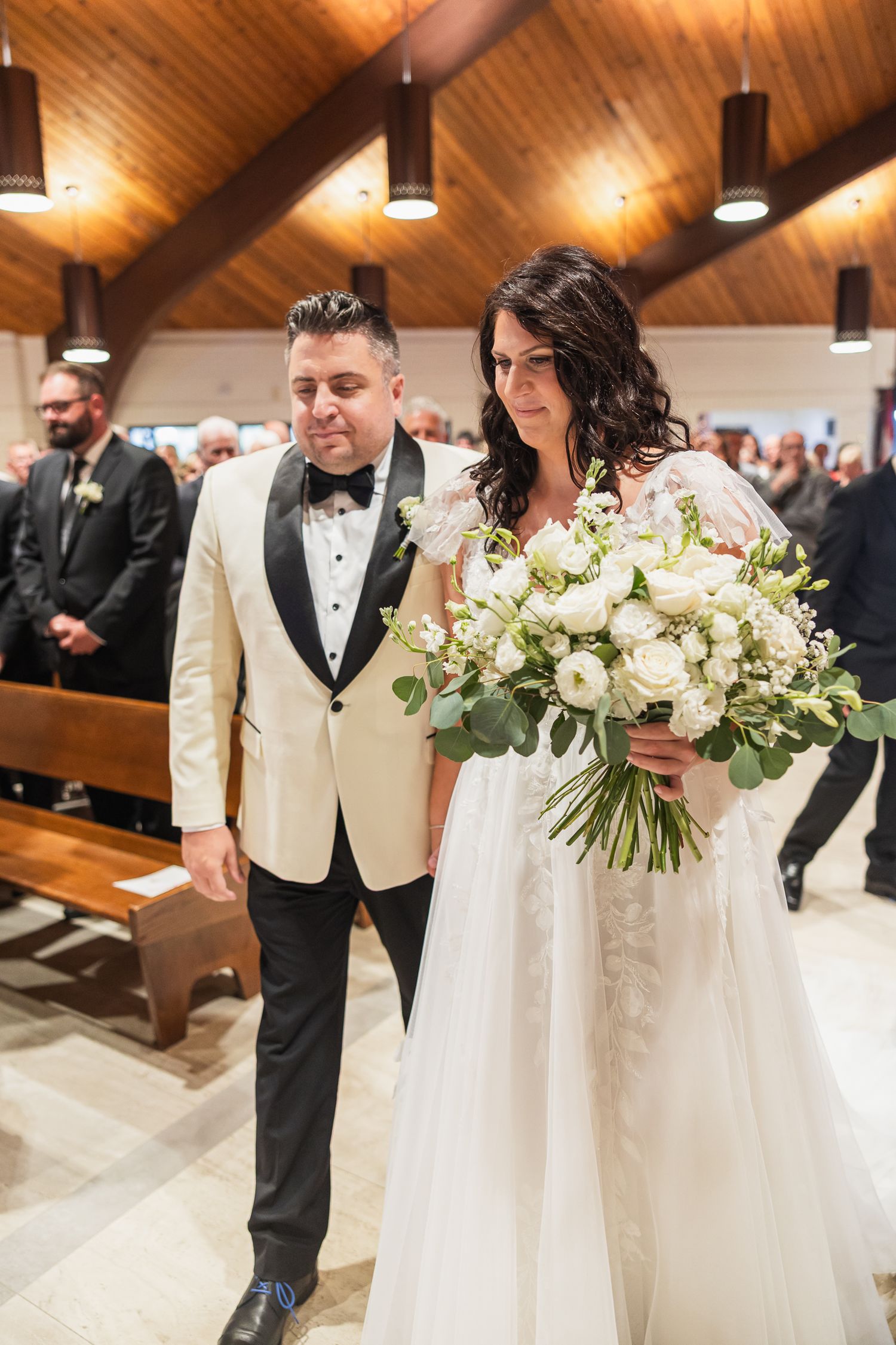 Bride holding white floral bouquet walks down church aisle with groom in cream tuxedo jacket.