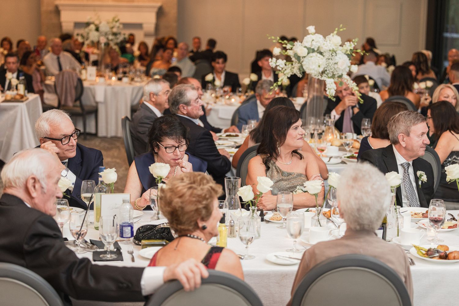 Elegant wedding reception with guests seated at round tables adorned with white floral centerpieces in a grand ballroom.