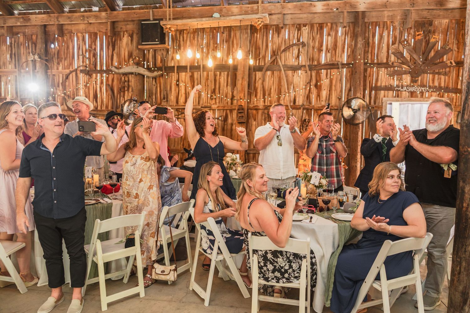 Wedding guests celebrate and raise glasses at rustic barn reception with wooden walls and string lights.