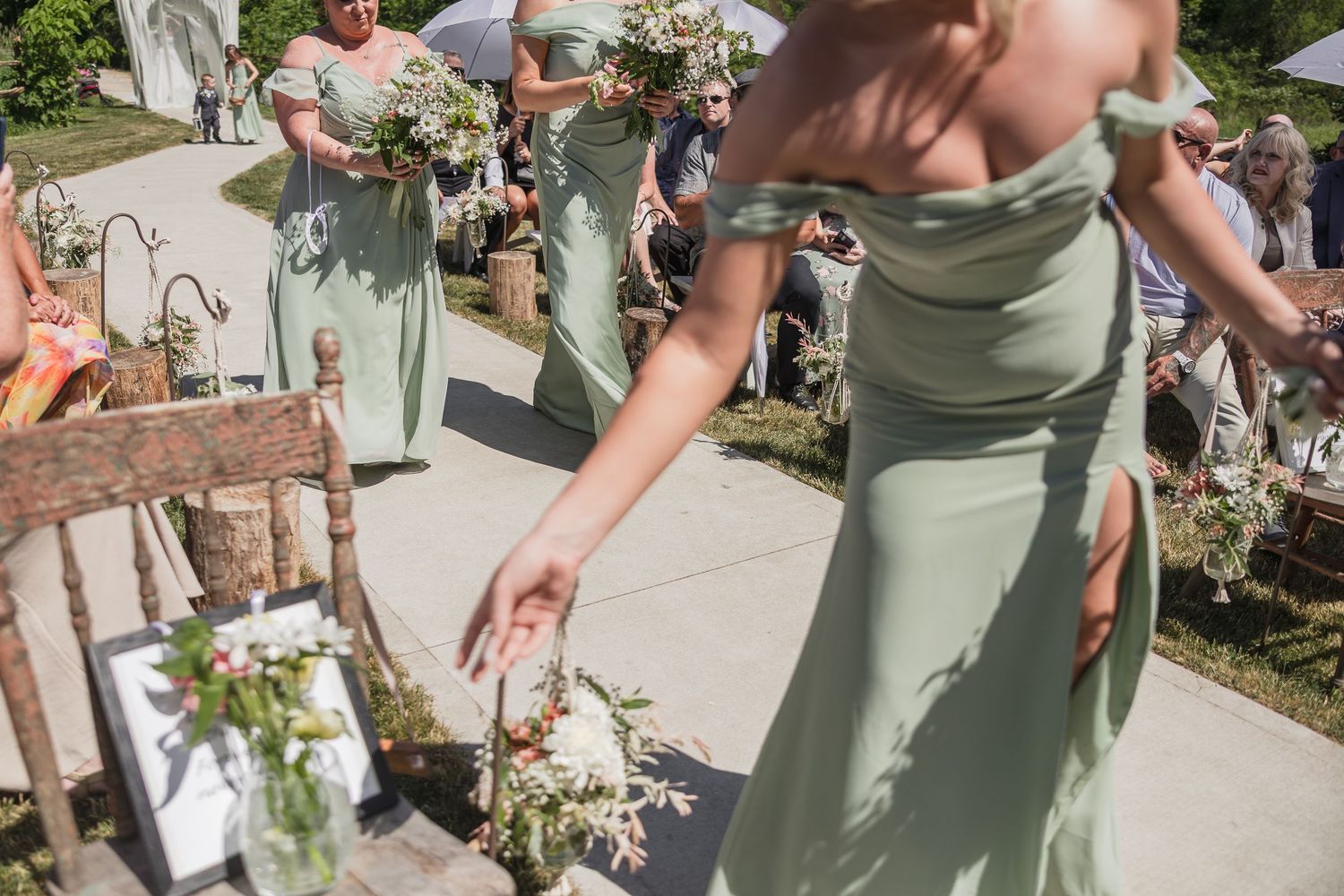 Bridesmaid in sage green off-shoulder dress scattering flower petals along outdoor wedding ceremony aisle.