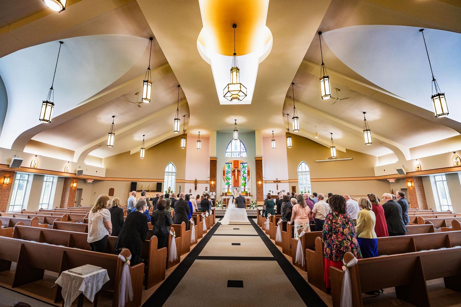 Wide angle view of church interior with guests seated in pews under vaulted ceiling and hanging lights.