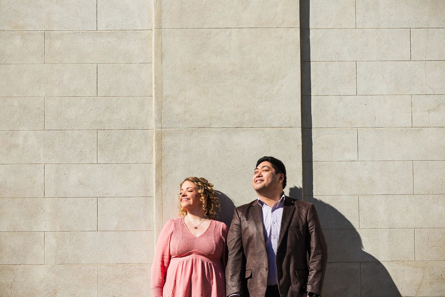 Couple laughing against white brick wall in sunlight during outdoor photoshoot.