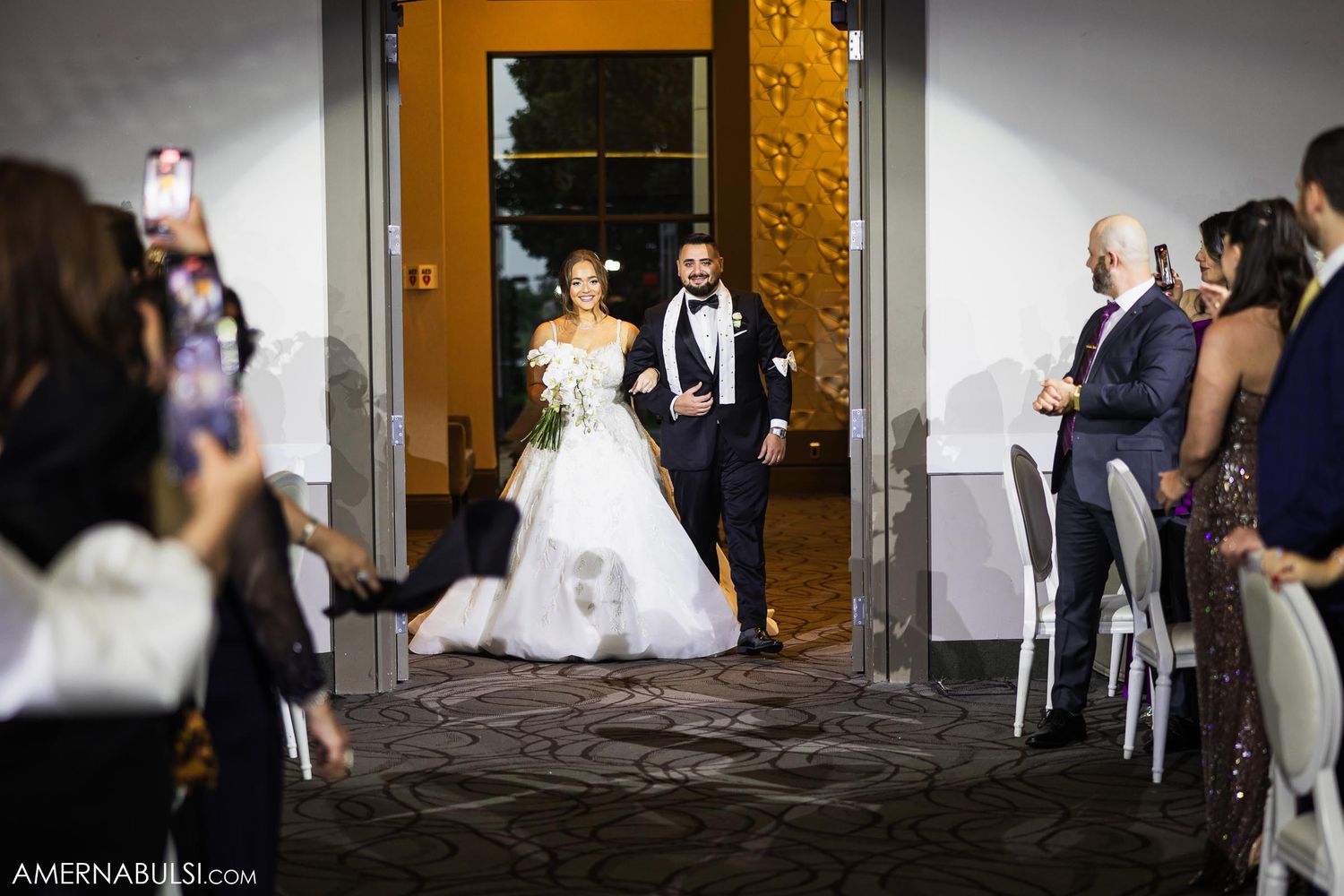A wedding couple makes their entrance through golden doors as guests look on from both sides.
