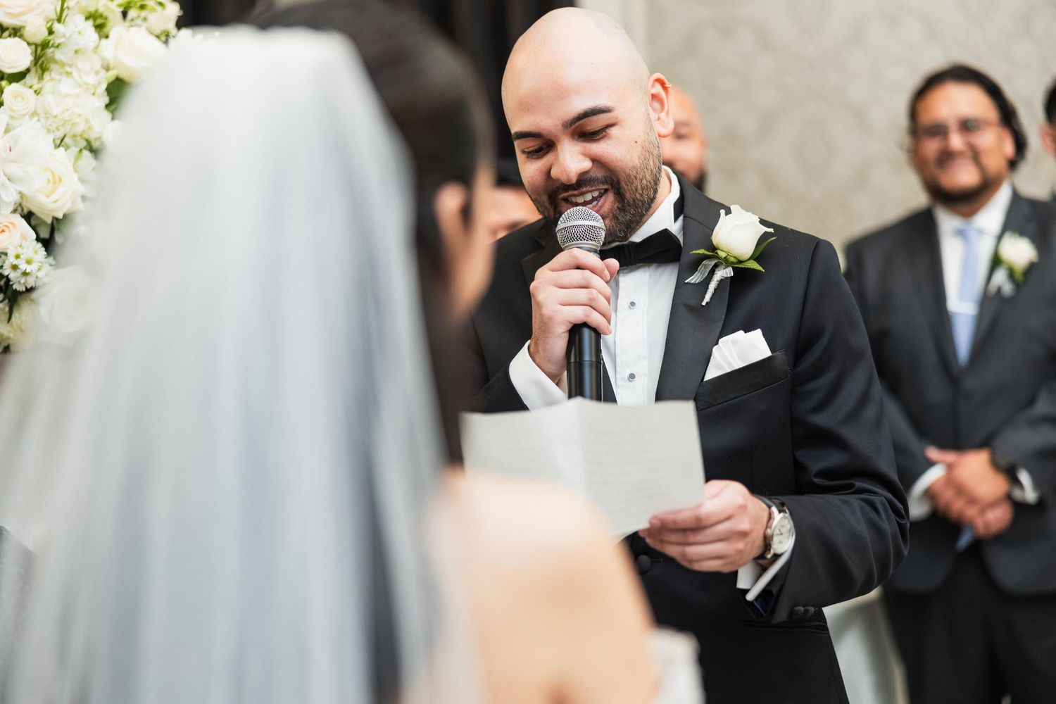 Dapper groom in black tuxedo reads wedding vows during emotional ceremony with white floral arrangements.