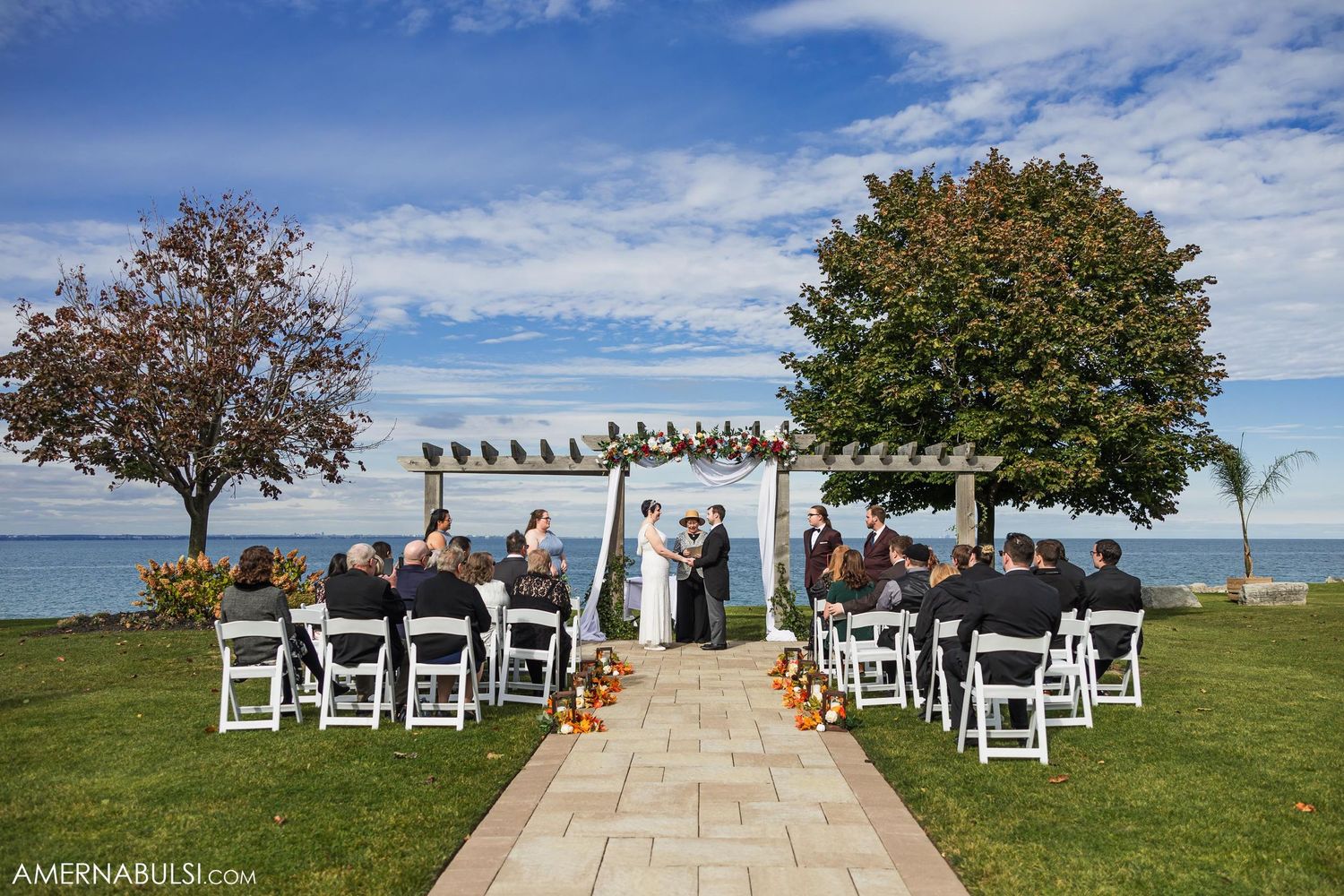 Intimate outdoor wedding ceremony setup with white chairs facing a pergola against waterfront backdrop.