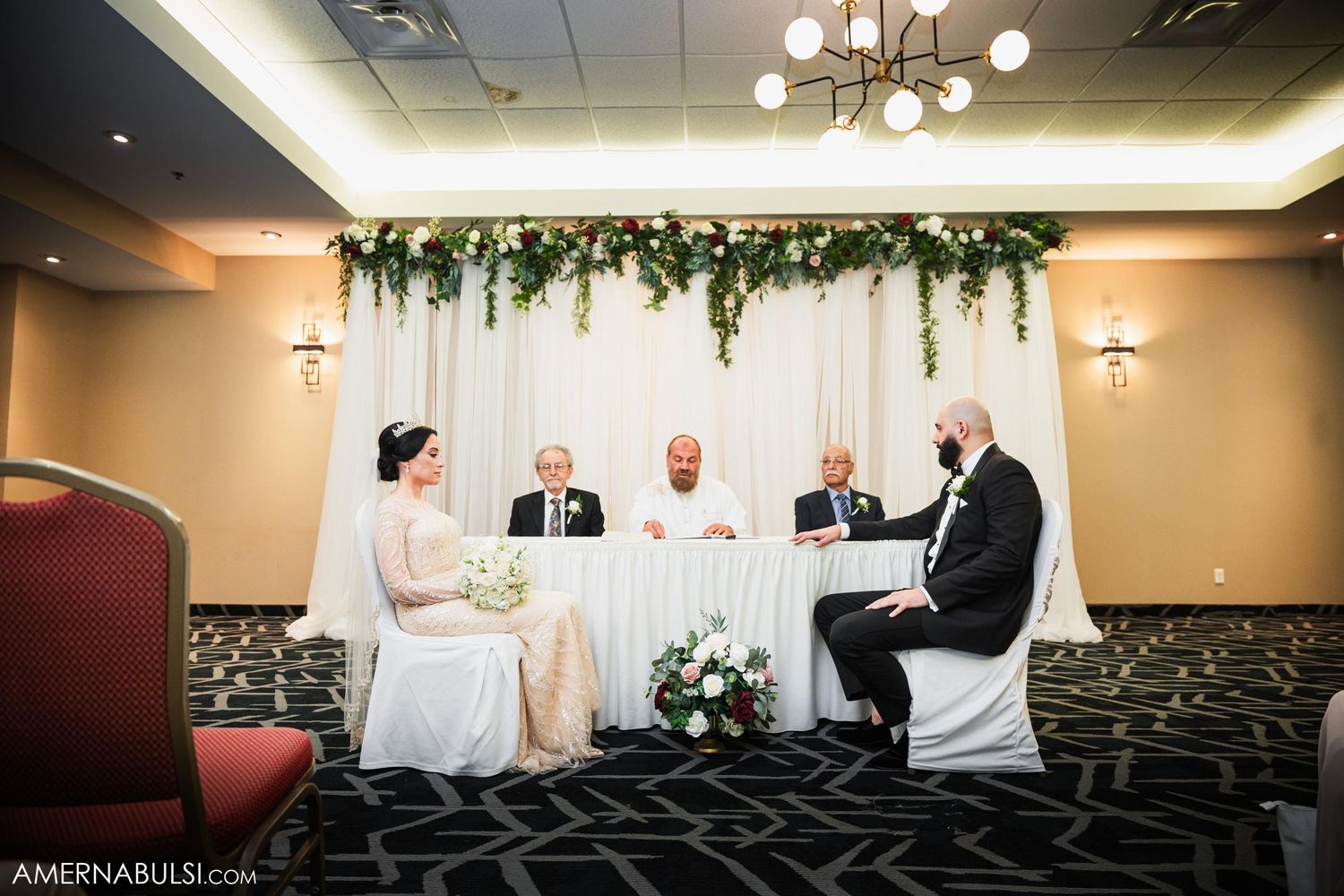 Wedding ceremony setup with floral arch and white backdrop in an elegant indoor venue.