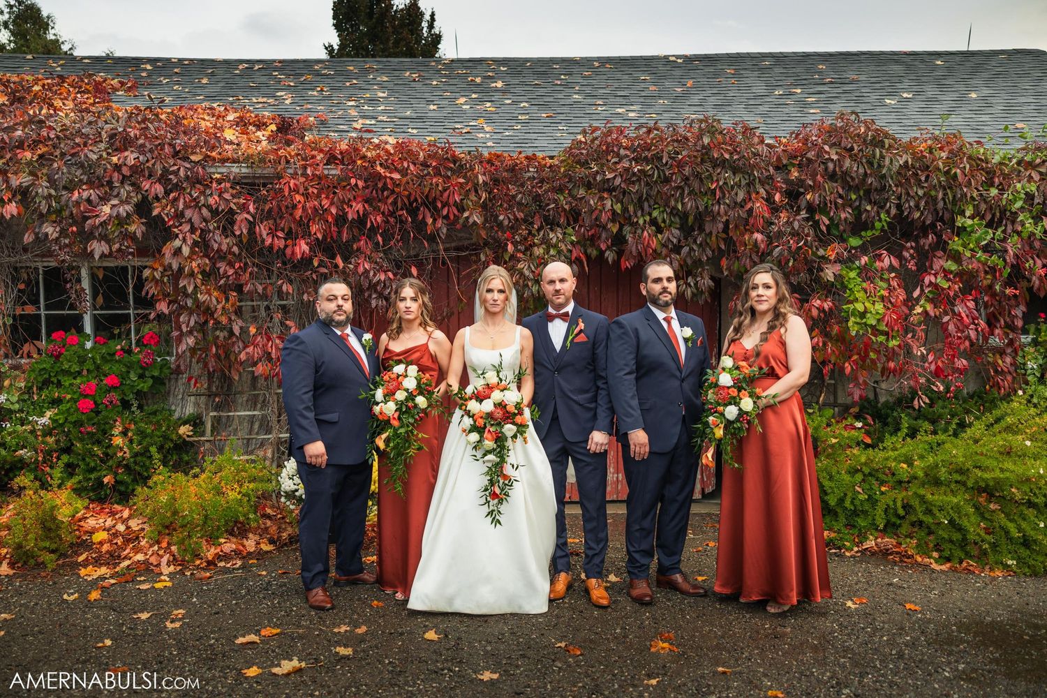 Wedding party poses in front of rustic barn with autumn foliage and burgundy accents.