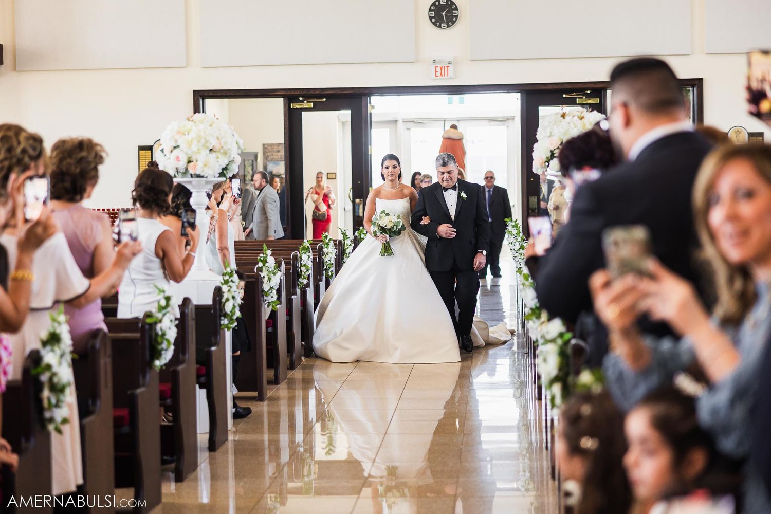 Guests capture photos on phones as the wedding couple walks down the church aisle.
