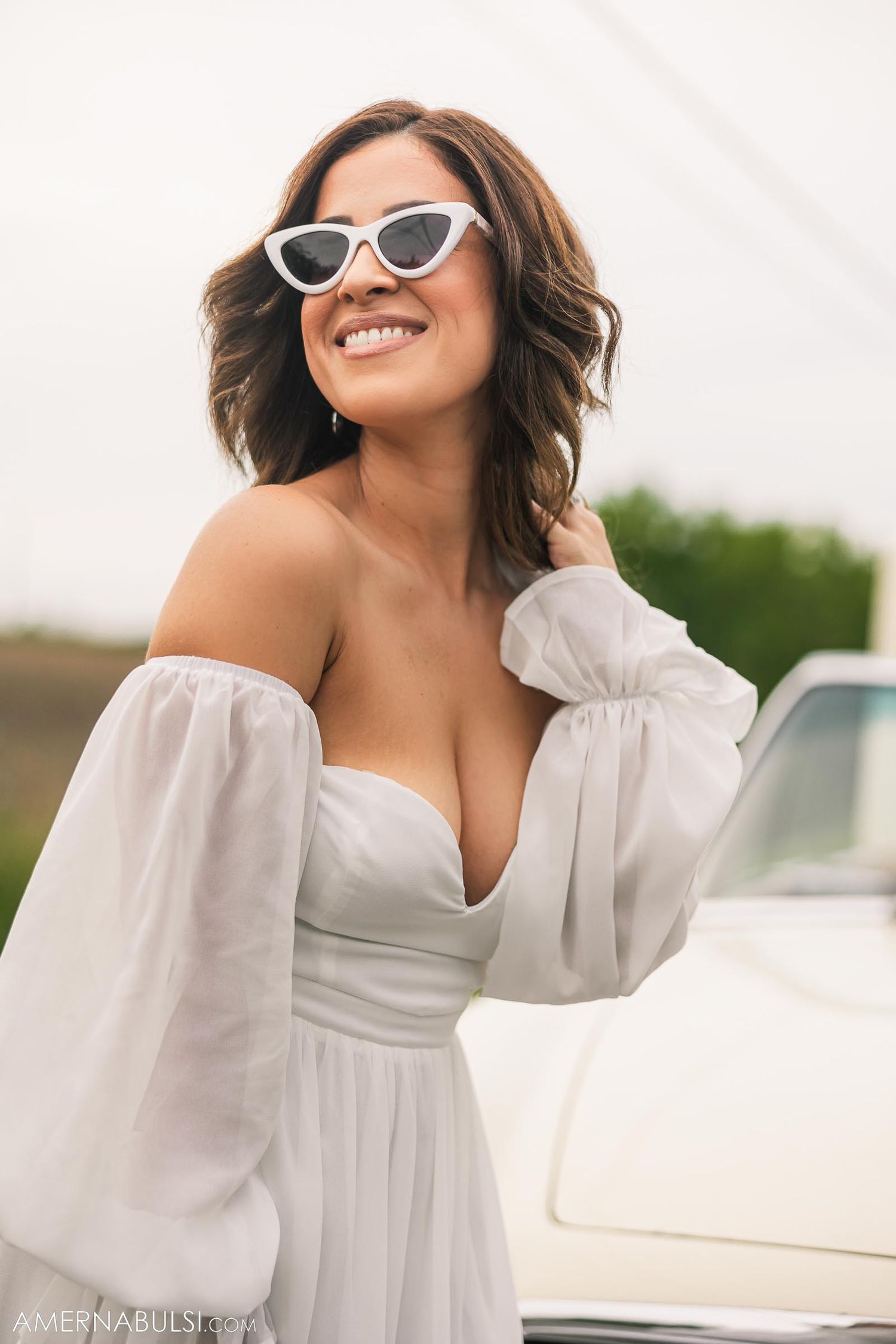 Woman in white off-shoulder dress and retro sunglasses poses next to classic car.