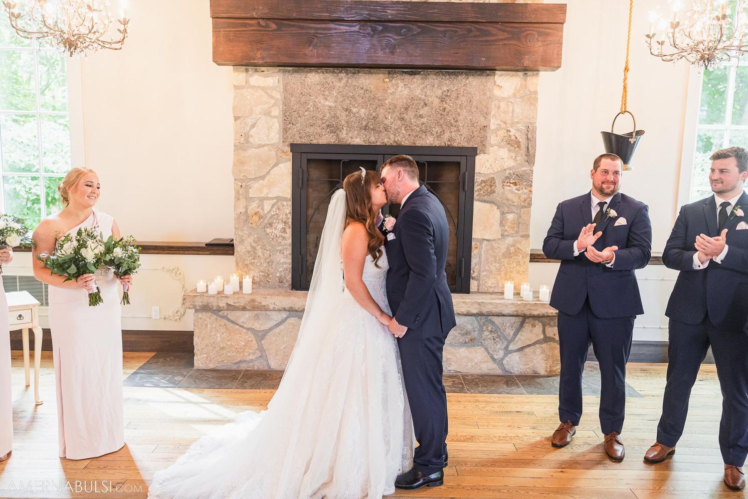 Wedding ceremony first kiss with guests applauding in elegant room with stone fireplace.