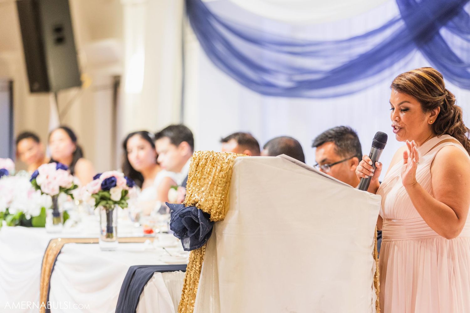 A bridesmaid in a peach dress gives an emotional wedding speech at an elegant reception with blue drapery.
