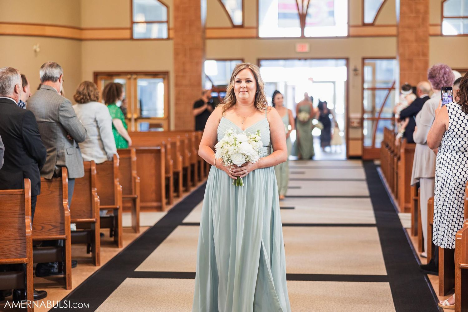 A wedding attendant in a pale blue gown walks down a church aisle carrying a white bouquet.