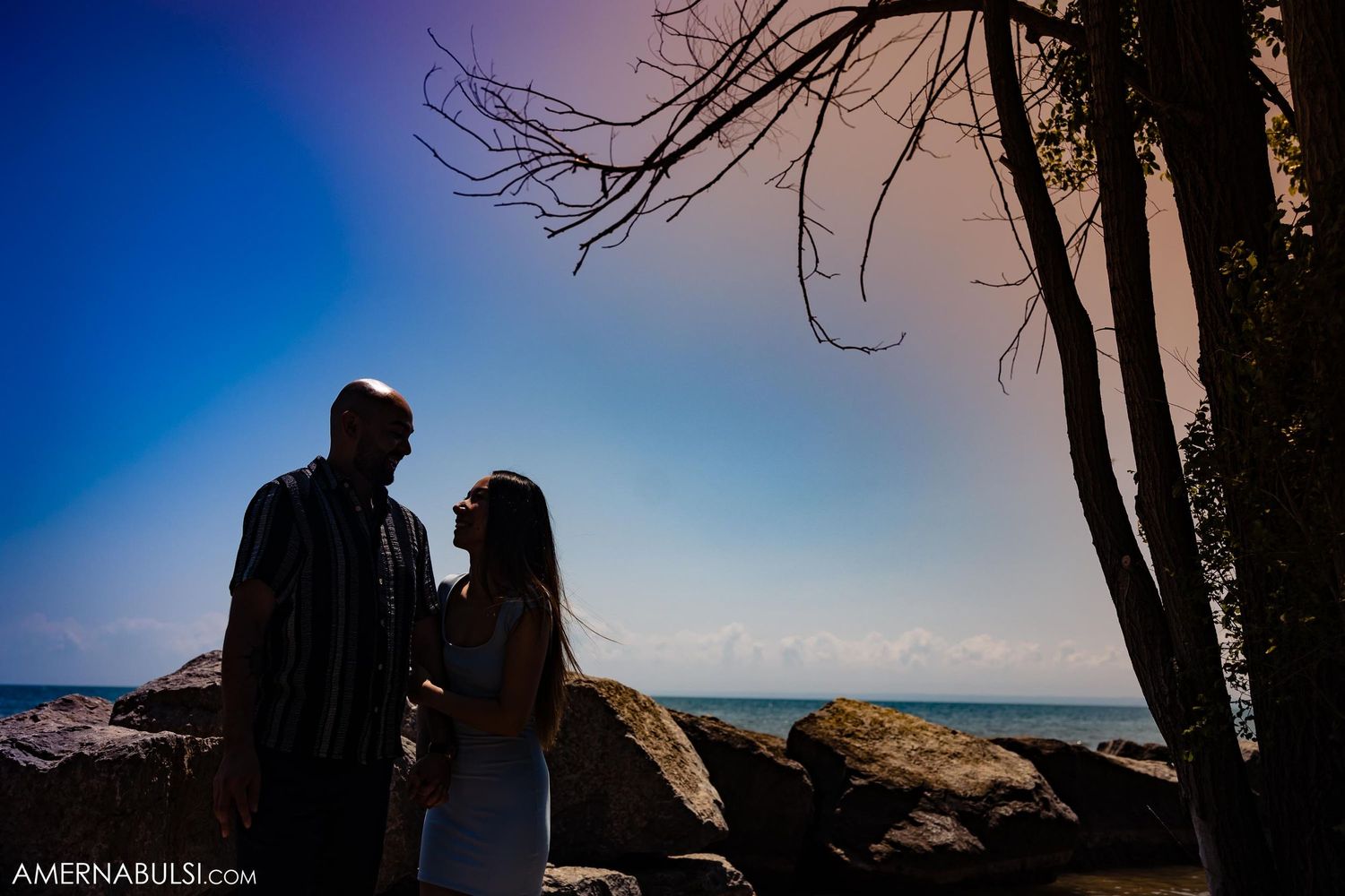 Silhouetted couple standing together against dramatic blue sky and ocean backdrop.