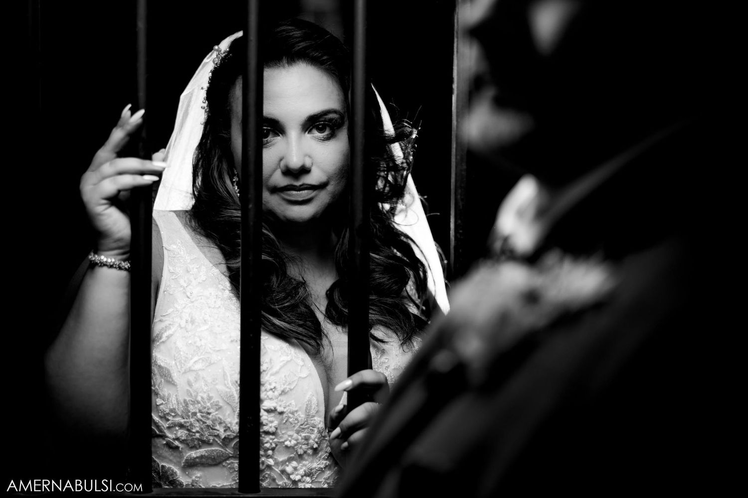 Black and white portrait of a bride in a lace dress looking through decorative metal bars.