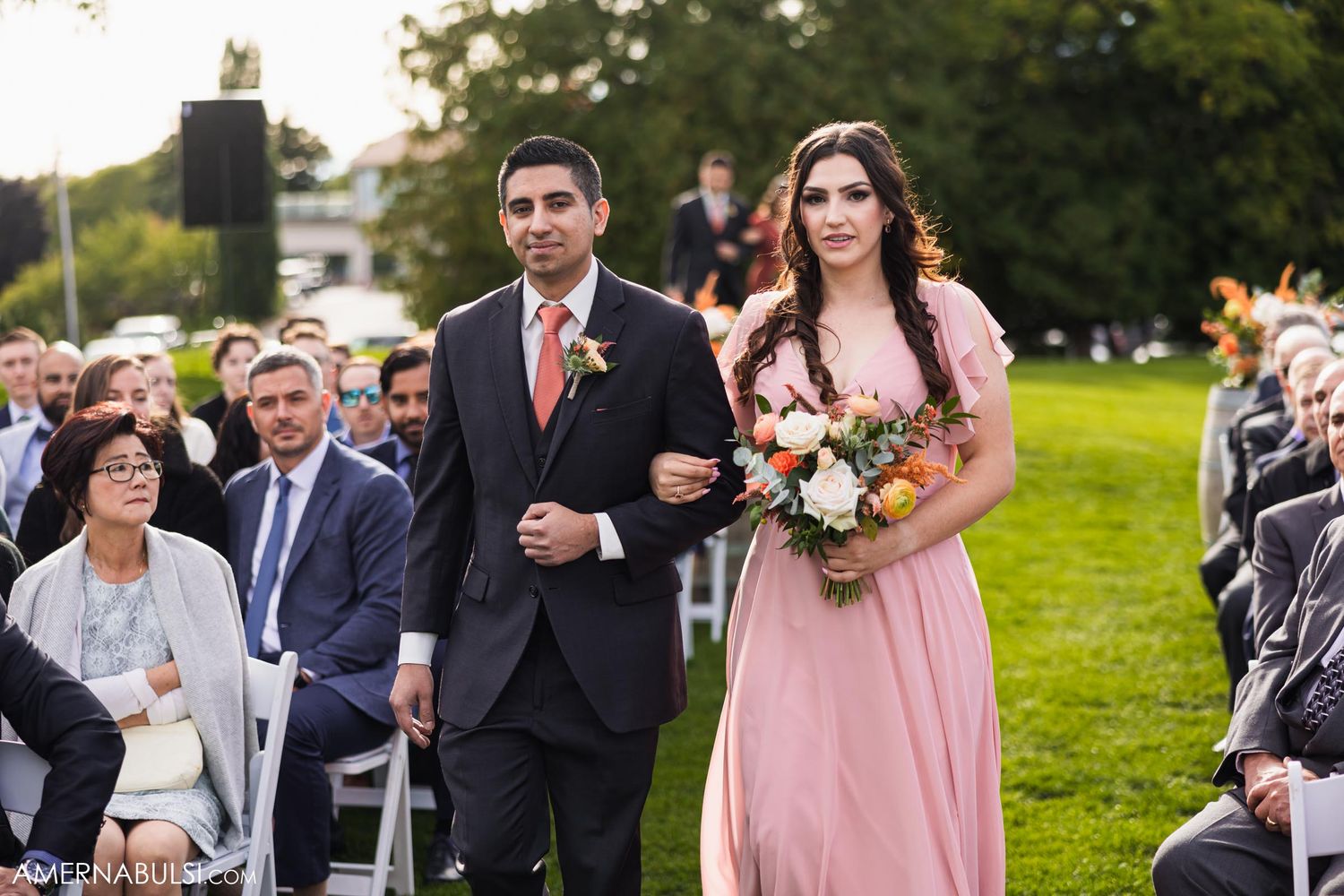 A bridesmaid in pink walks down the aisle with a groomsman during a wedding processional.
