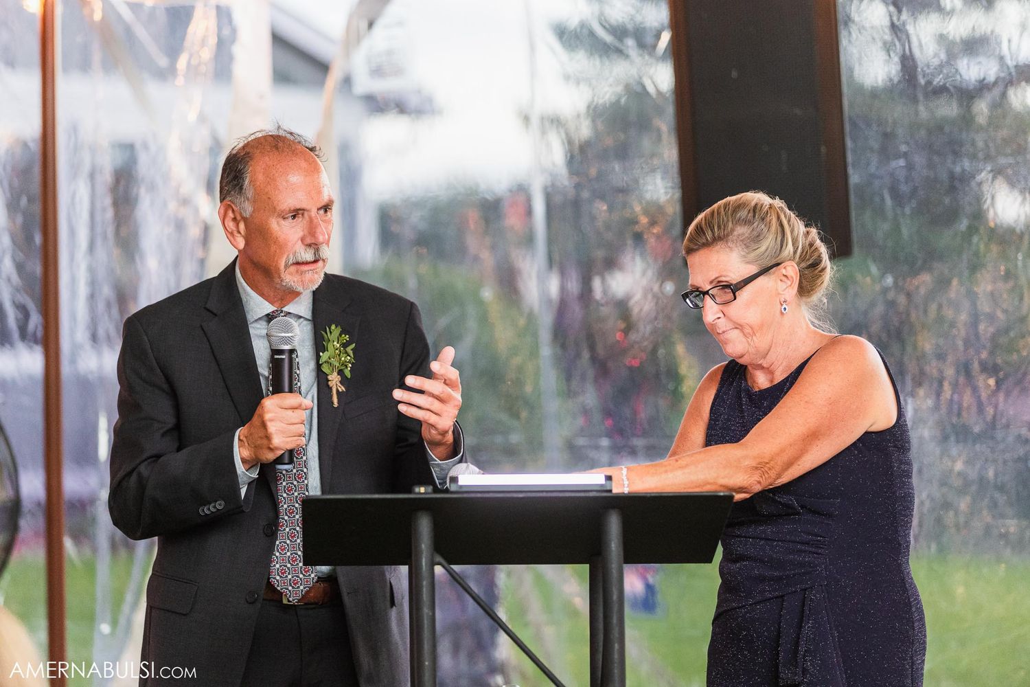 Two speakers share remarks at a wedding reception podium during evening hours.