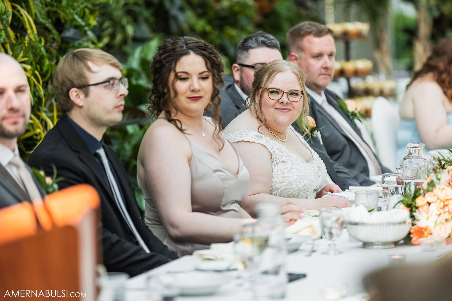 Wedding guests seated at a long reception table with floral centerpieces and greenery backdrop.