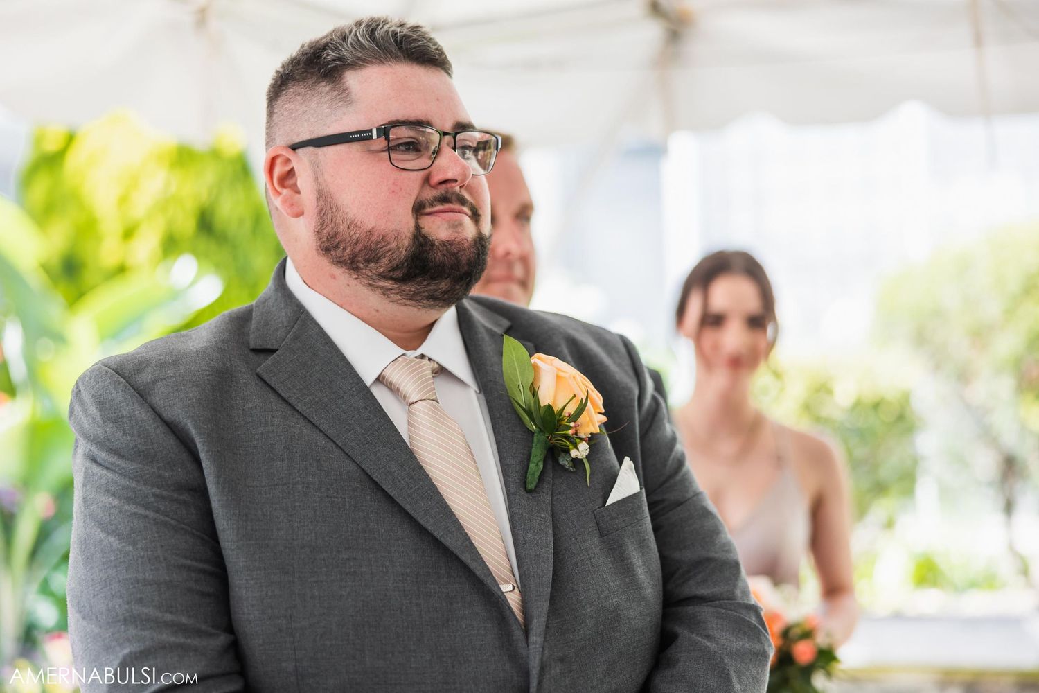 A formal wedding ceremony under a white tent with a participant wearing a grey suit and yellow boutonniere.