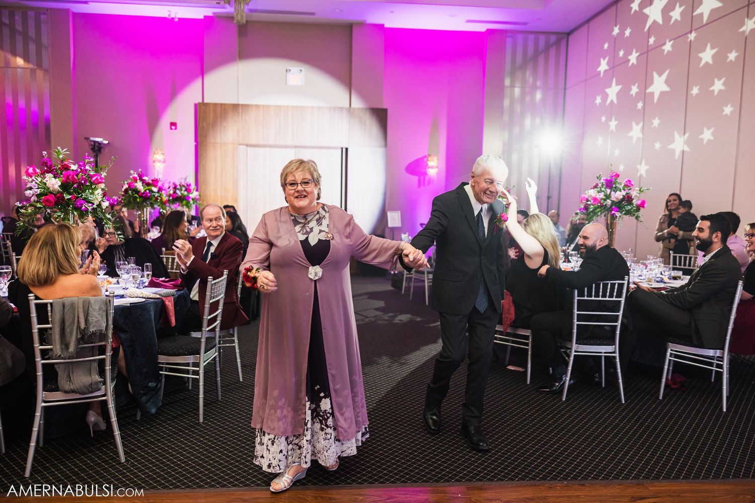 Guests dancing and celebrating at wedding reception with pink wall lighting and round dining tables.