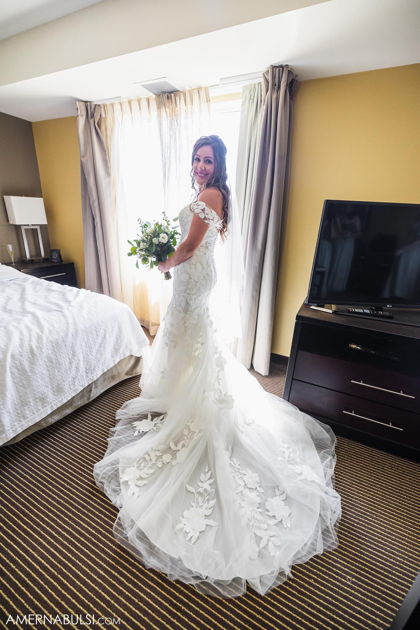 A bride in a lace wedding gown with long train poses in front of a window in a hotel room.