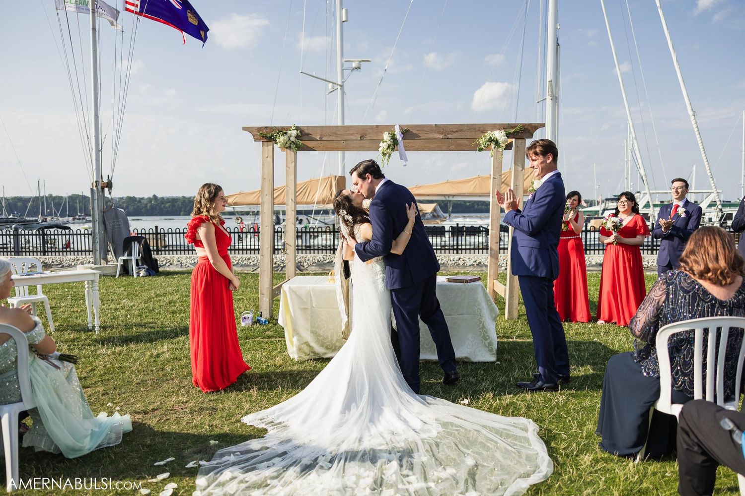 Waterfront wedding ceremony with attendants in red dresses and white floral arch.