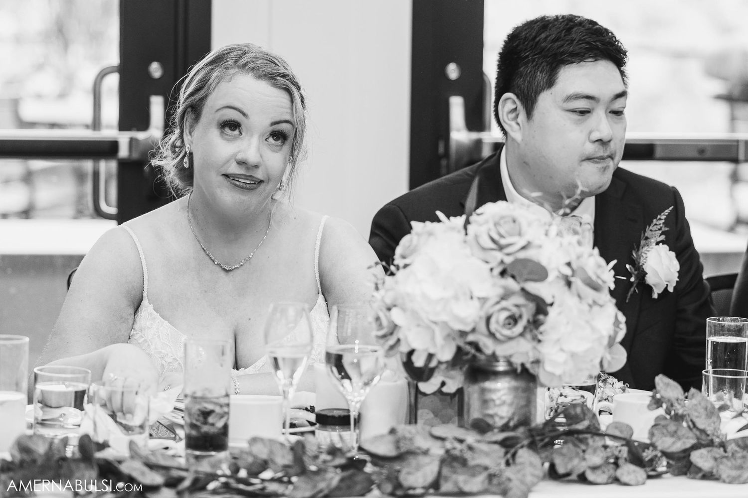 A couple sits at their wedding reception head table surrounded by elegant floral centerpieces.