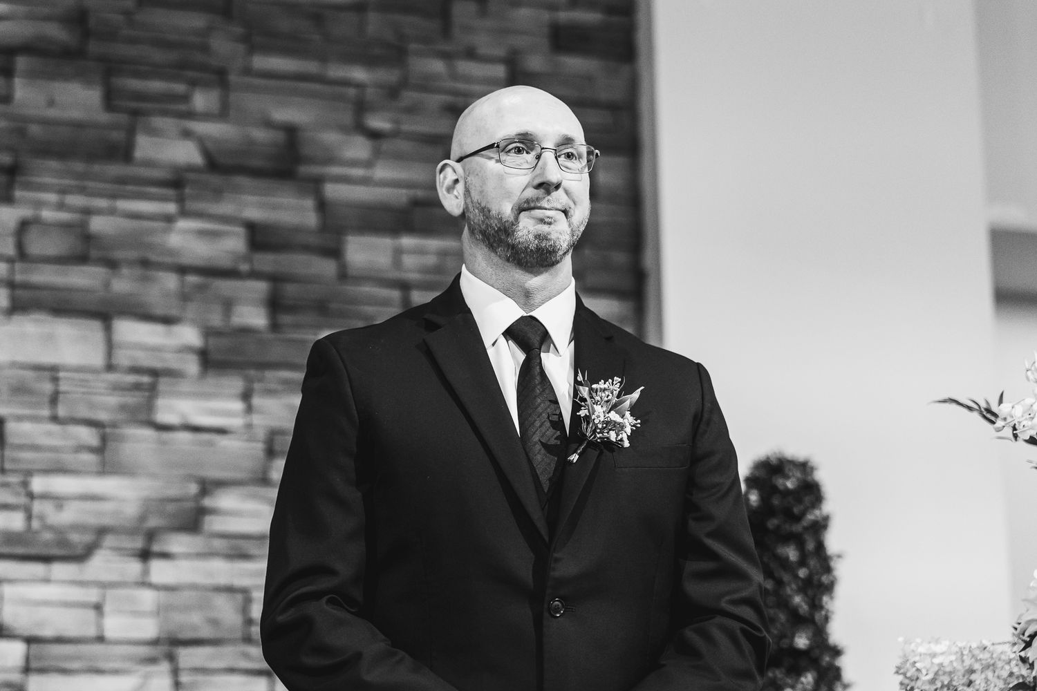 Black and white portrait of a wedding attendant wearing a dark suit against a stone wall.