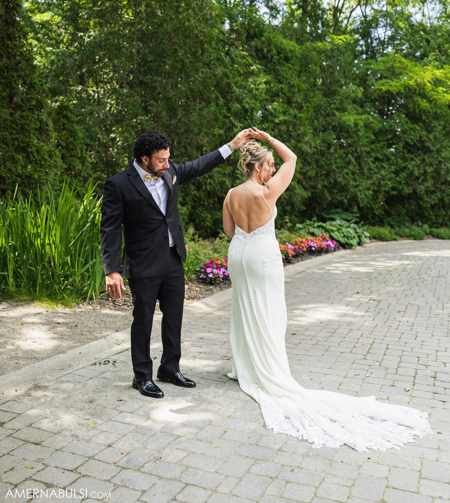 Bride in flowing white gown twirls on stone pathway while groom watches.