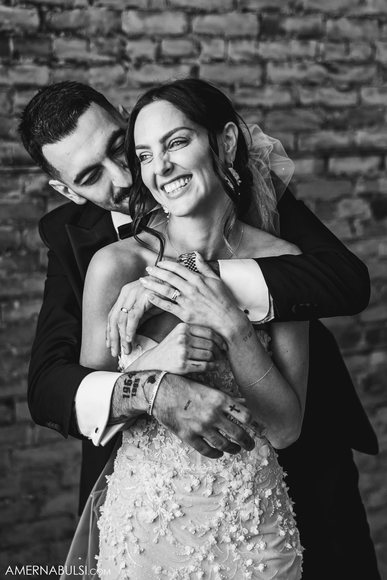 Black and white portrait of newlyweds sharing a candid laugh against brick wall.