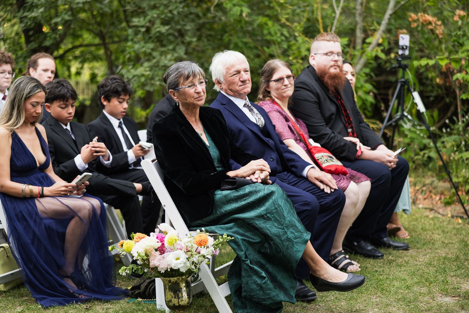 Wedding guests seated in white chairs watch an outdoor ceremony surrounded by trees and flowers.